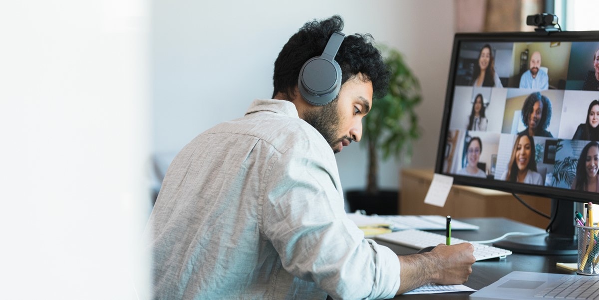 man in a virtual meeting from home wearing headphones, writing at desk