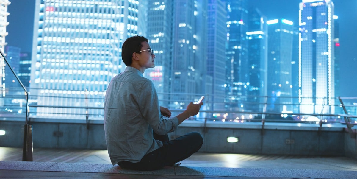 man sitting on building rooftop, lit up skyline in front of him