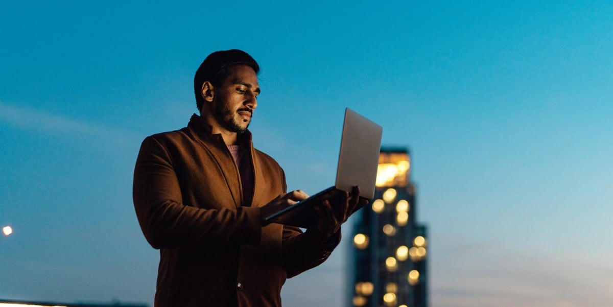professional man holding laptop, standing outside, skyscraper behind him