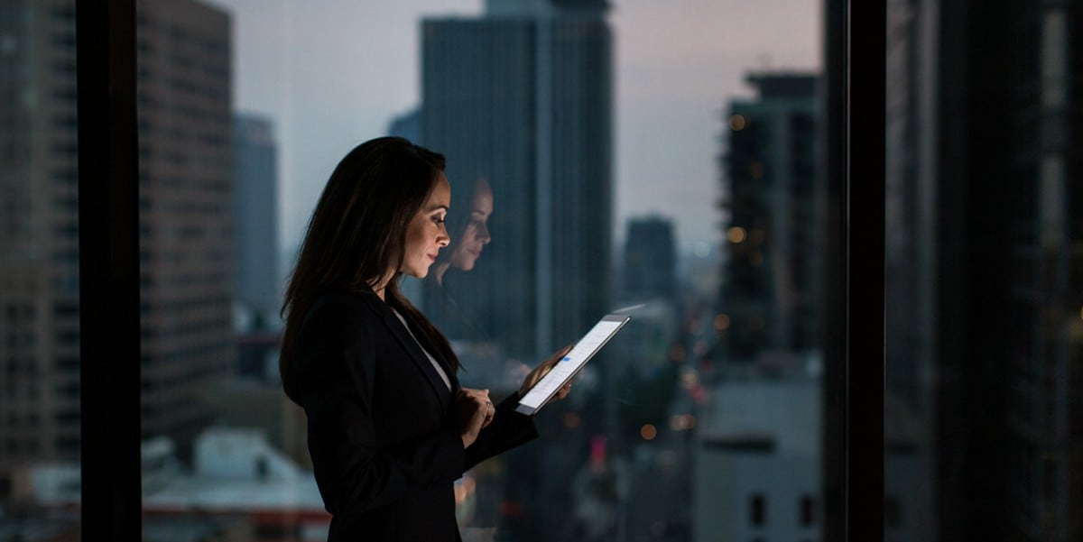 professional woman using tablet, standing in front of window with skyscrapers in the background