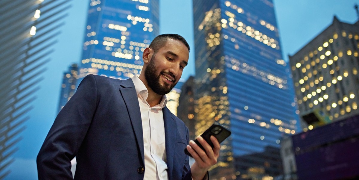 professional man standing outside in front of skyscrapers, looking down at cellphone