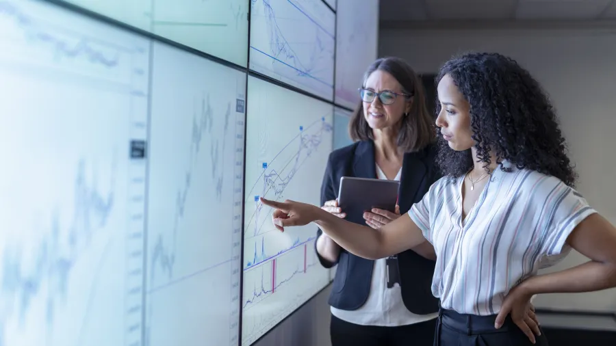 One woman pointing at giant screen and another woman with clipboard next to her